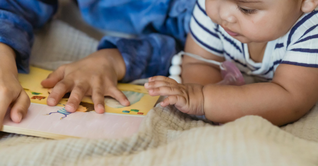 A baby and another child lie on their tummies, looking at a board book together.