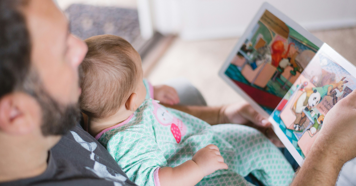 A father reads a board book to a baby. The dad can be seen from an upper angle. He is holding a baby in his lap and has a book with a colorful illustration open in his hands. Both the baby and father are looking at the book.